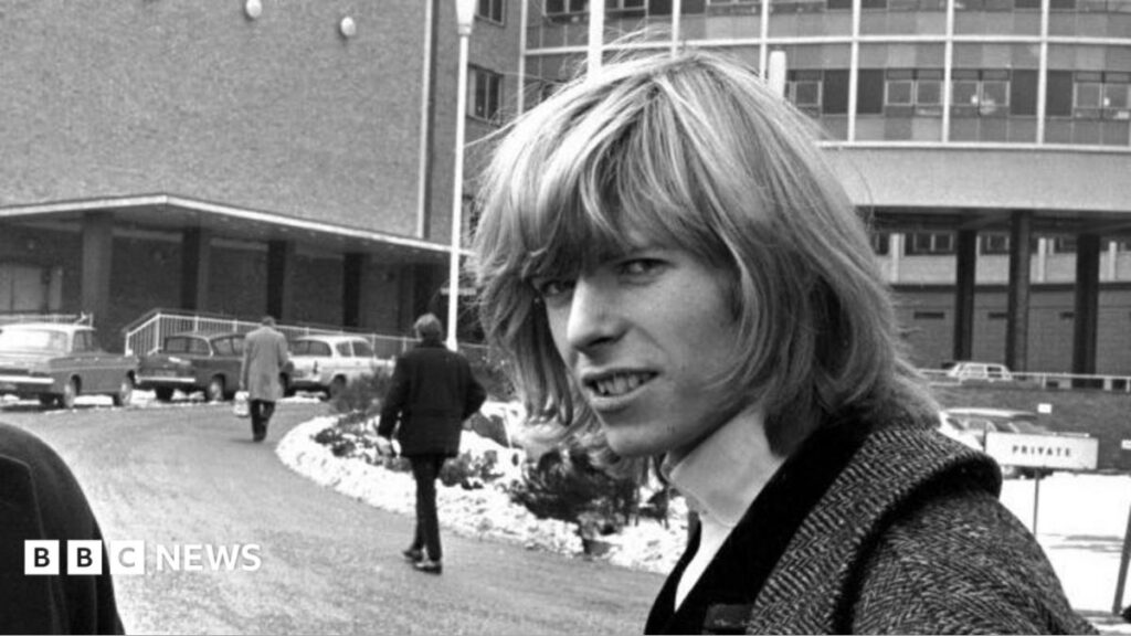 Lost Bowie tracks from 1960s released for first time Earl Spencer walking, wearing a navy suit and a purple tie.