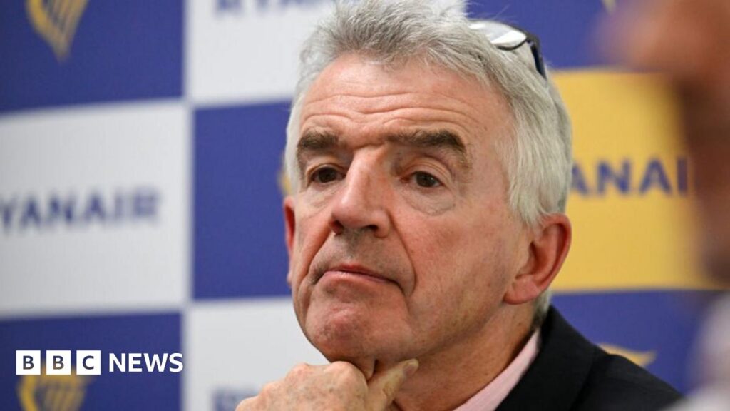 Michael O'Leary, who has short, grey hair and wears a black jacket over a pink shirt, sits at a table with his hand resting on his chin. A board with Ryanair logos are on a board behind him.