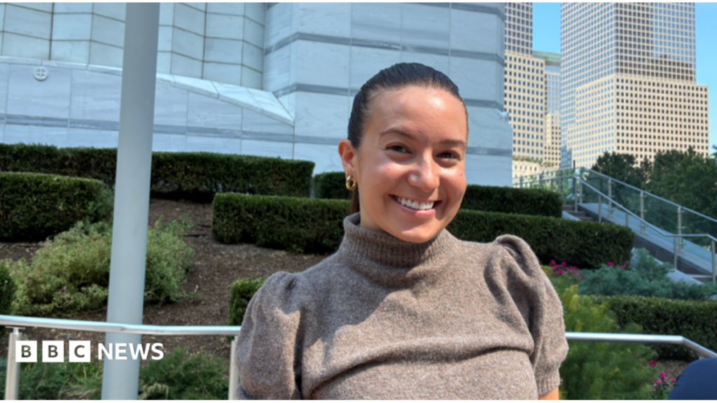 How soon into dating do you talk about your salary? A woman, with brown hair tied back, smiles while standing in a park. She wears a short sleeved grey top and gold earrings