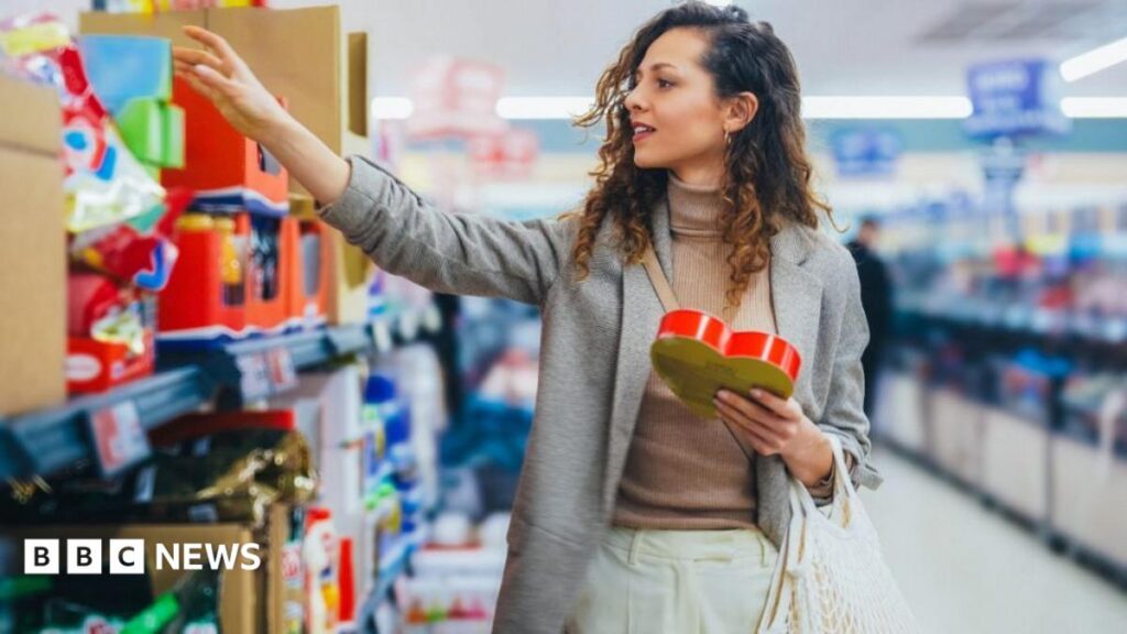 What are the new rules in Scotland’s supermarkets? A woman compares items on a supermarket shelf. She has long, brown hair and is wearing a grey jacket over a brown jumper and cream trousers.